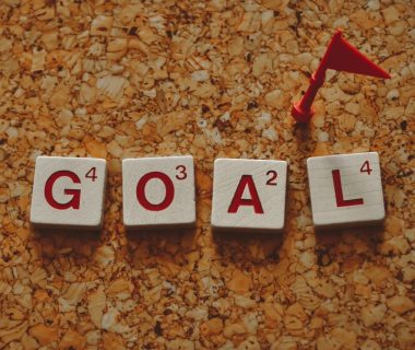 Scrabble tiles forming the word 'goal' with a small red flag on a cork board background.